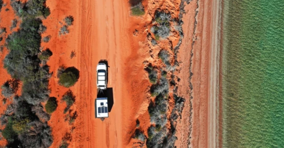 Aerial view of a vehicle towing a caravan along a red dirt outback road bordered by green vegetation
